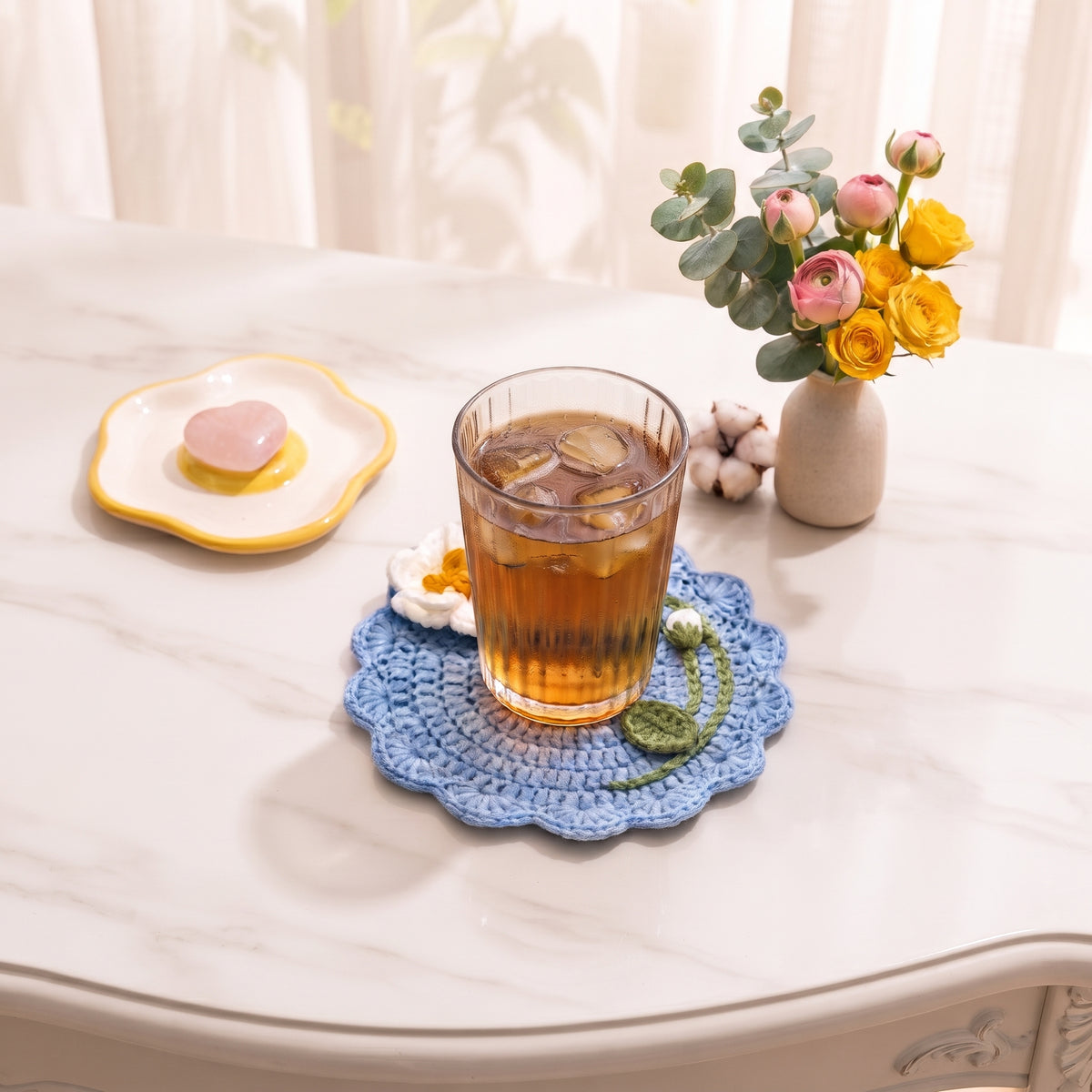 Blue handmade crochet flower coaster under tall glass of iced tea on white marble table, small rose bouquet and cotton stems, elegant aesthetic table setting