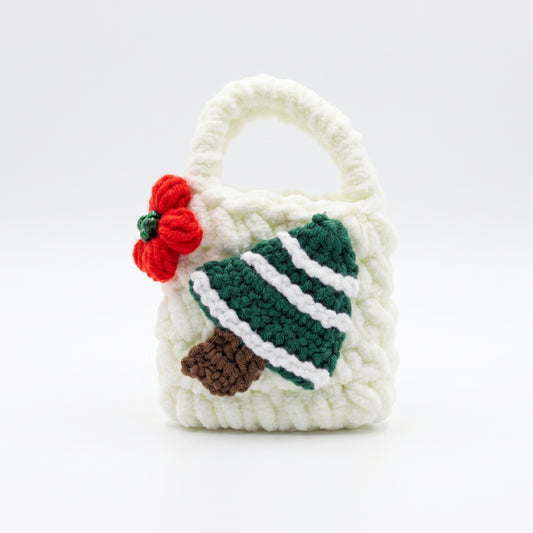 A close-up view of a small, white crochet earbud pouch. It features a handwoven green Christmas tree with white stripes and a little red knit flower with green beads. The bag has a tiny loop handle and is set against a plain white background, perfect for holiday-themed tech accessories.