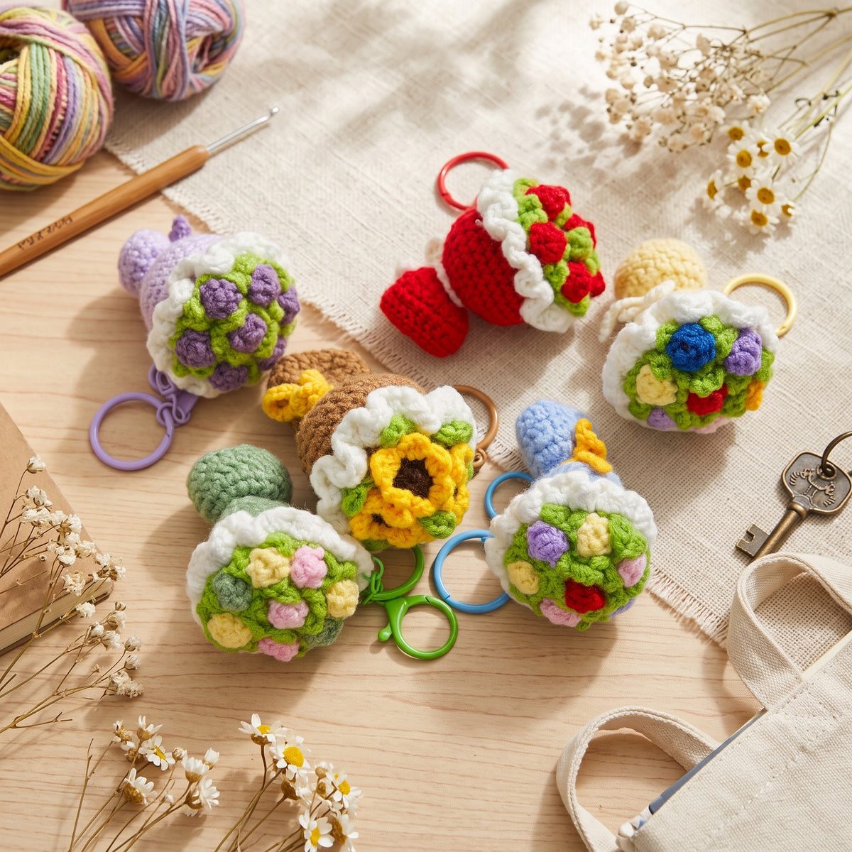 Assorted handmade crochet flower bouquet keychains laid on linen table with yarn, crochet hook and dried daisies, handmade knitted key accessories