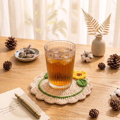 Handmade beige crochet coaster with two 3D yellow sunflowers and green leaves, placed under glass of water on cozy warm desk with table lamp and tulip vase, cute boho table decor