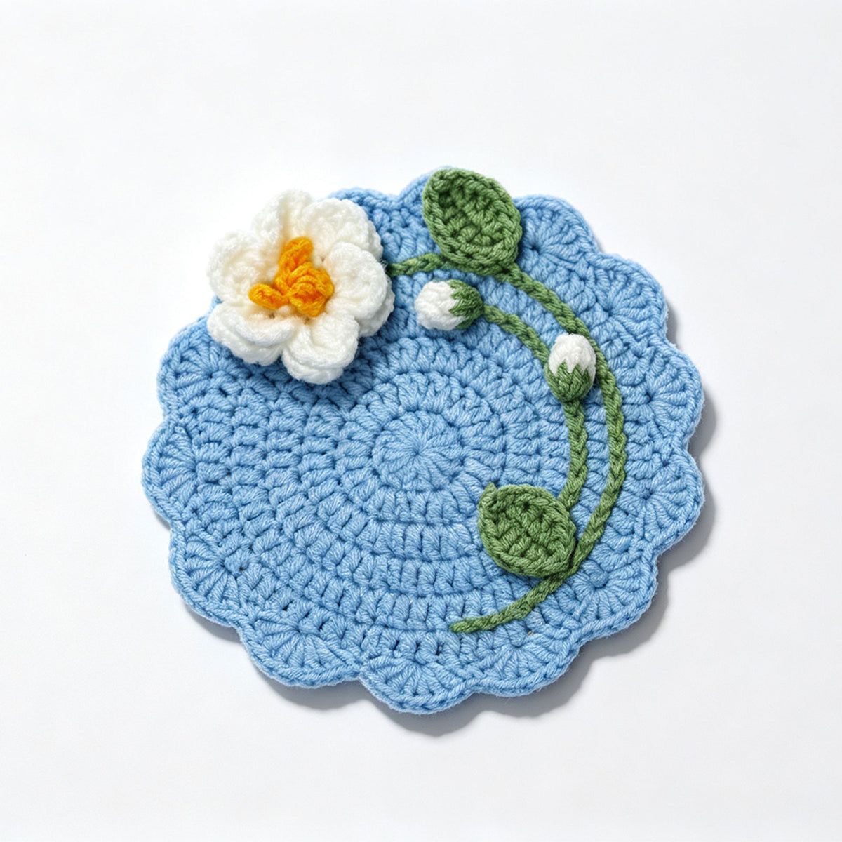 Blue handmade crochet flower coaster with white blossom, green leaves and bud, scalloped edge, flat lay on plain white background, floral drink mat