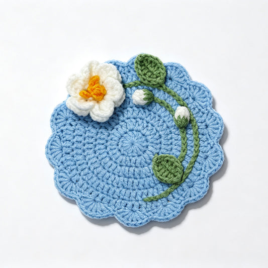 Blue handmade crochet flower coaster with white blossom, green leaves and bud, scalloped edge, flat lay on plain white background, floral drink mat