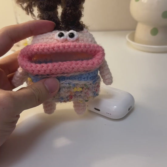 Hands inserting a white AirPods Pro case into a funny handmade crochet monster pouch with wild brown hair and big pink lips.