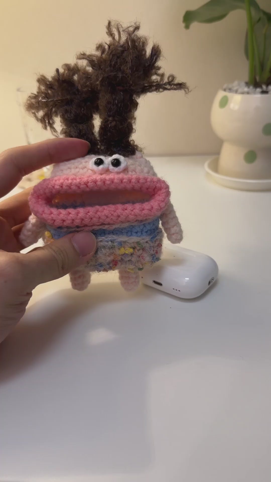 Hands inserting a white AirPods Pro case into a funny handmade crochet monster pouch with wild brown hair and big pink lips.